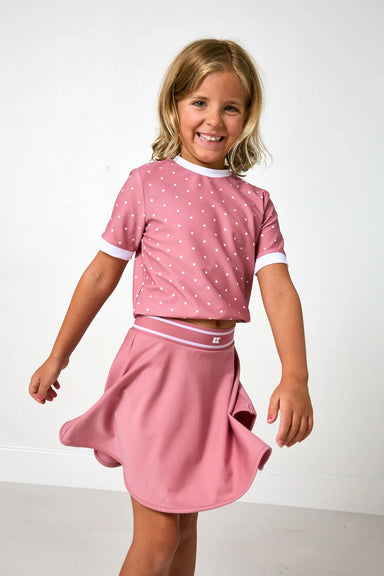 A young girl smiling and twirling while wearing a pink two-piece outfit consisting of a short-sleeve crop top with white polka dots and white trim, and a matching pink skirt with a white waistband.