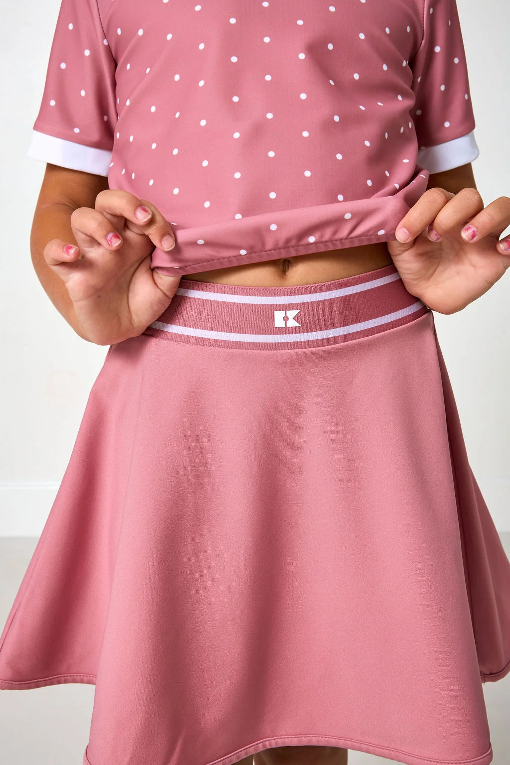A close-up of a child wearing a dusty pink two-piece outfit with a white polka-dot short-sleeve top and a matching flared skirt with a white and pink waistband. 