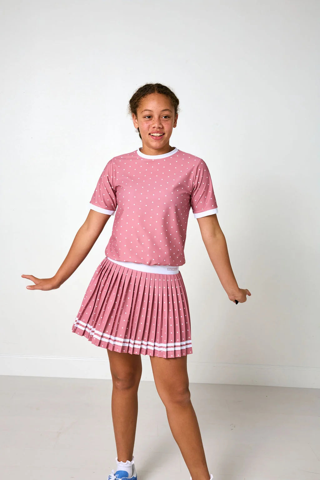 A young girl modeling a pink and white two-piece outfit consisting of a short-sleeve t-shirt with white dots and a matching pleated skirt with white stripes, set against a plain white studio background.