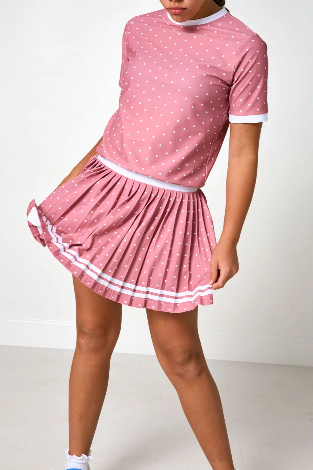 A pink short-sleeve top and pleated mini skirt set with white polka dots and white trim, shown on a model against a plain white studio background.
