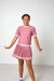 A young girl modeling a pink and white two-piece outfit consisting of a short-sleeve t-shirt with white dots and a matching pleated skirt with white stripes, set against a plain white studio background.