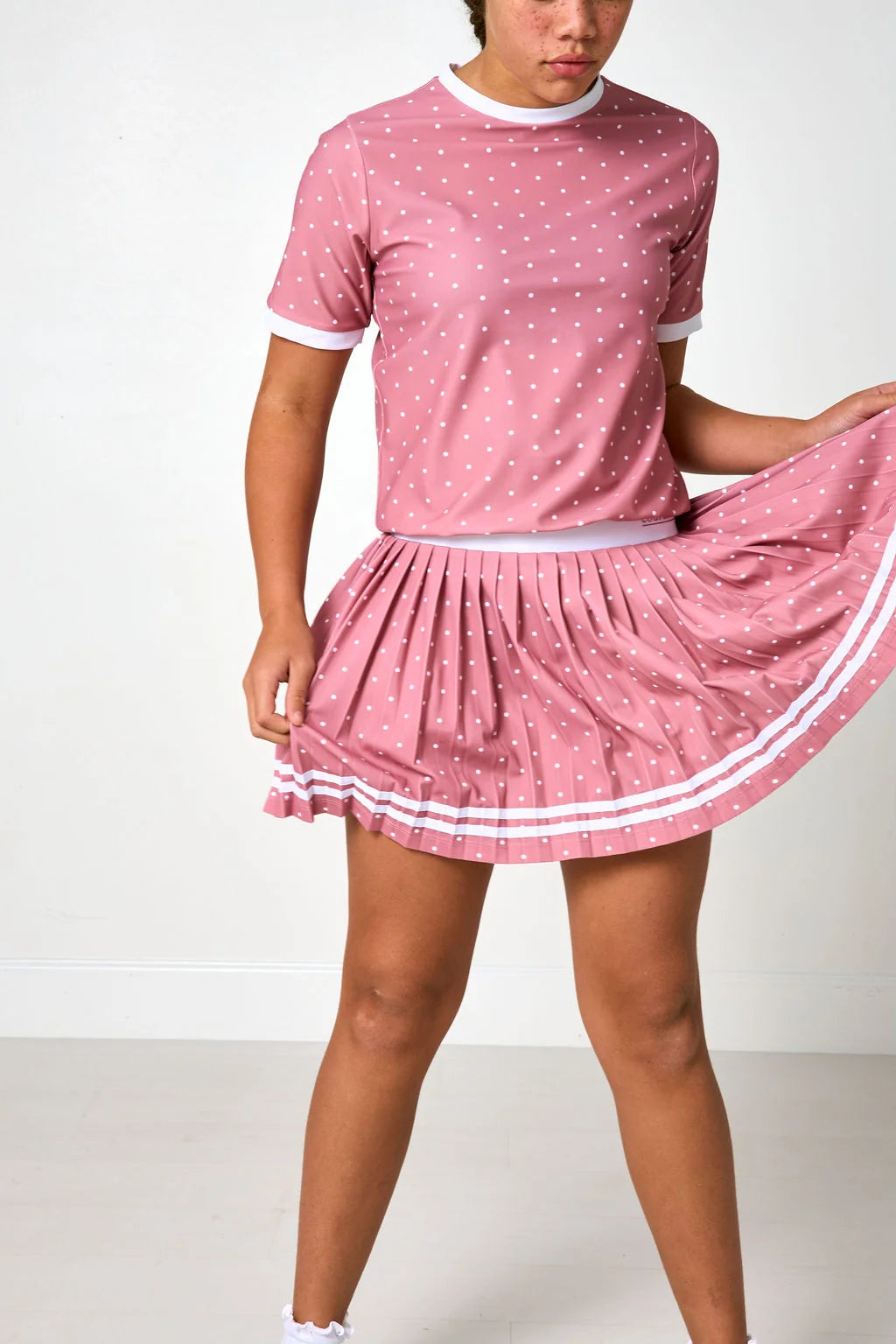 A model wearing a pink short-sleeve t-shirt and matching pleated skirt set with white polka dots and white trim, shown against a plain white studio background.