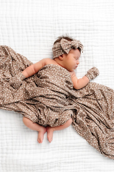 A baby covered in a square blanket, a matching bow headband, and a pair of mittens, all in a brown and white ditsy floral print on a solid white background.