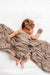 A baby covered in a square blanket, a matching bow headband, and a pair of mittens, all in a brown and white ditsy floral print on a solid white background.