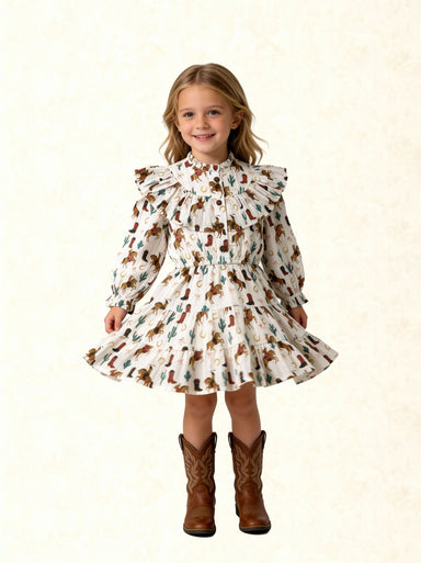 A young girl with blonde hair stands against a light beige textured background wearing a long-sleeve white dress with a brown and green western print, featuring a ruffled collar and a flared skirt, paired with brown cowboy boots.