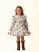 A young girl with blonde hair stands against a light beige textured background wearing a long-sleeve white dress with a brown and green western print, featuring a ruffled collar and a flared skirt, paired with brown cowboy boots.