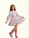 A young girl with long brown hair is wearing a light pink long-sleeve dress with a brown and gold floral pattern, paired with pink cowboy boots, against a textured cream background.