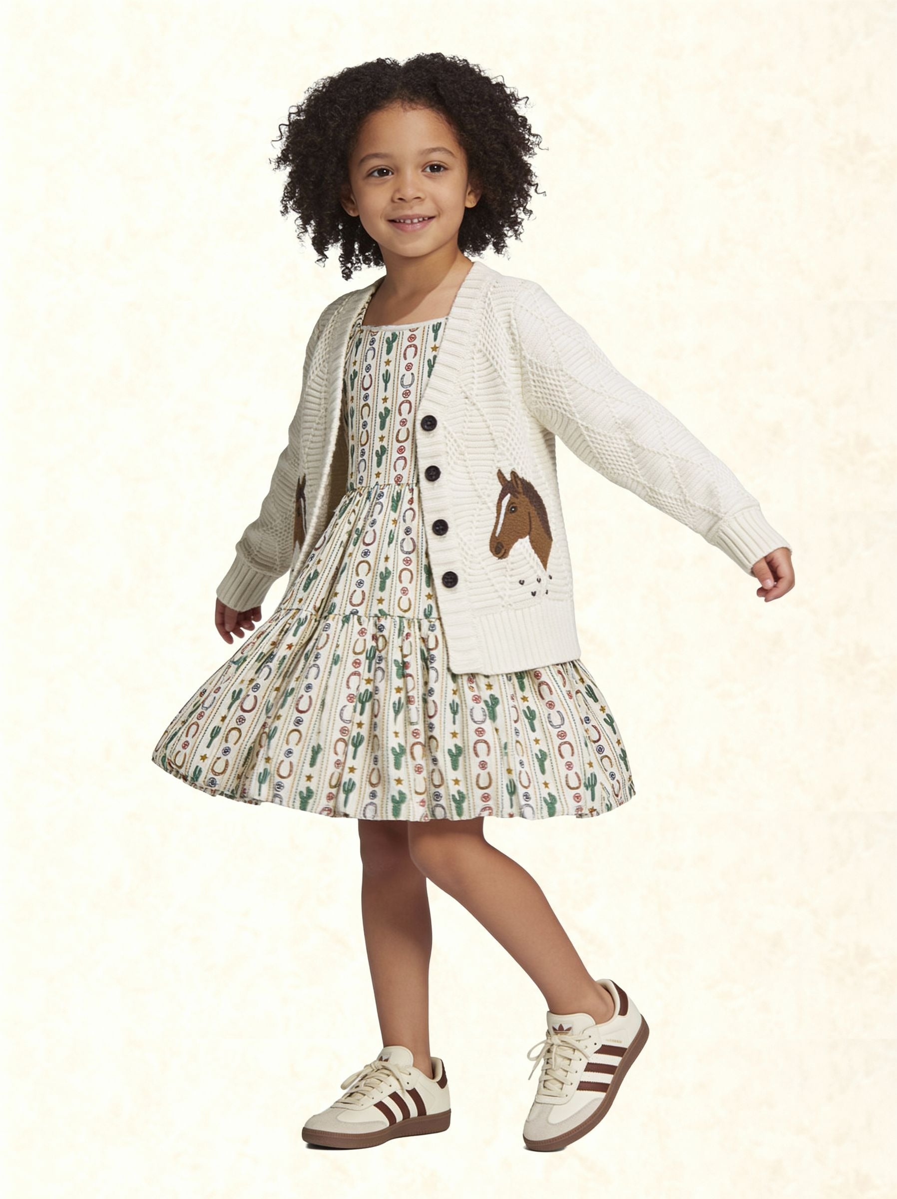 A young girl with curly dark hair is wearing a white knit cardigan with a brown horse patch and black buttons, paired with a matching patterned dress and white sneakers with brown stripes, set against a light cream textured background.