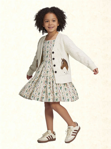 A young girl with curly dark hair is wearing a white knit cardigan with a brown horse patch and black buttons, paired with a matching patterned dress and white sneakers with brown stripes, set against a light cream textured background.