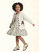 A young girl with curly dark hair is wearing a white knit cardigan with a brown horse patch and black buttons, paired with a matching patterned dress and white sneakers with brown stripes, set against a light cream textured background.