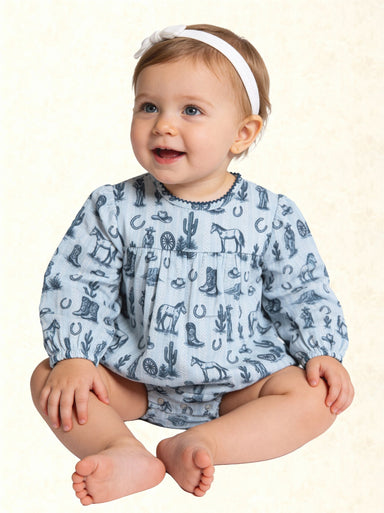 A baby girl sitting cross-legged wearing a light blue long-sleeve romper with a dark blue western-themed print of boots, horseshoes, cacti, and horses, paired with a white headband.