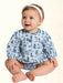 A baby girl sitting cross-legged wearing a light blue long-sleeve romper with a dark blue western-themed print of boots, horseshoes, cacti, and horses, paired with a white headband.