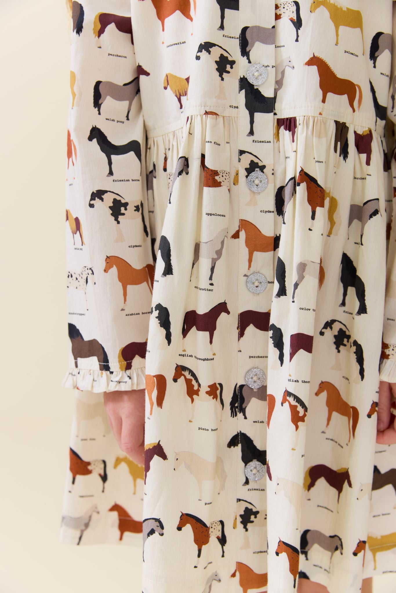 A close-up of a cream-colored dress with a whimsical all-over print of various horse illustrations in brown, black, white, and tan. The dress features tiered ruffles at the waist and cuffs, with a small portion of a model's hand visible on the left side.