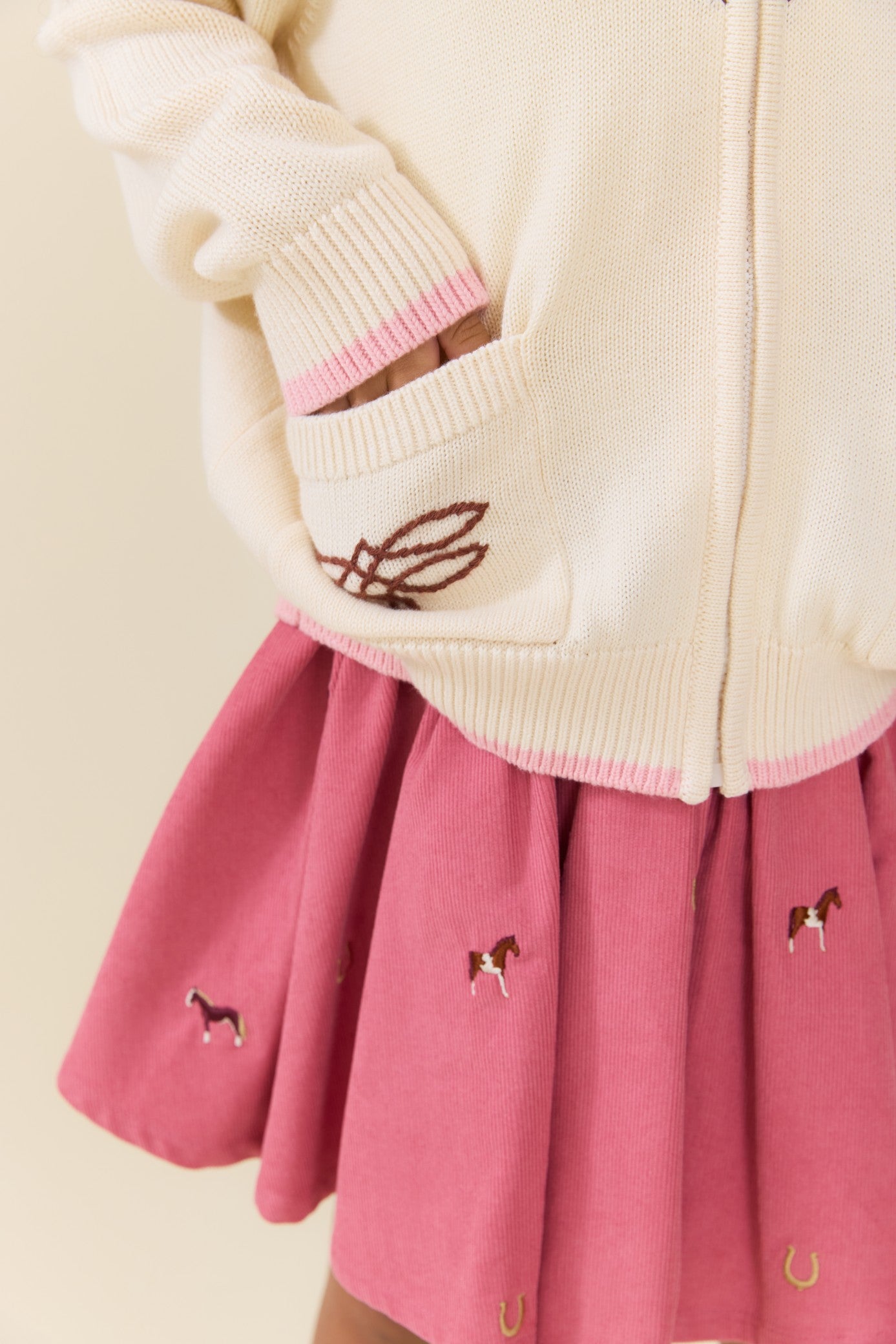 Pink pleated skirt with embroidered horses worn with a cream knit cardigan featuring a front pocket and pink trim by Pink Chicken