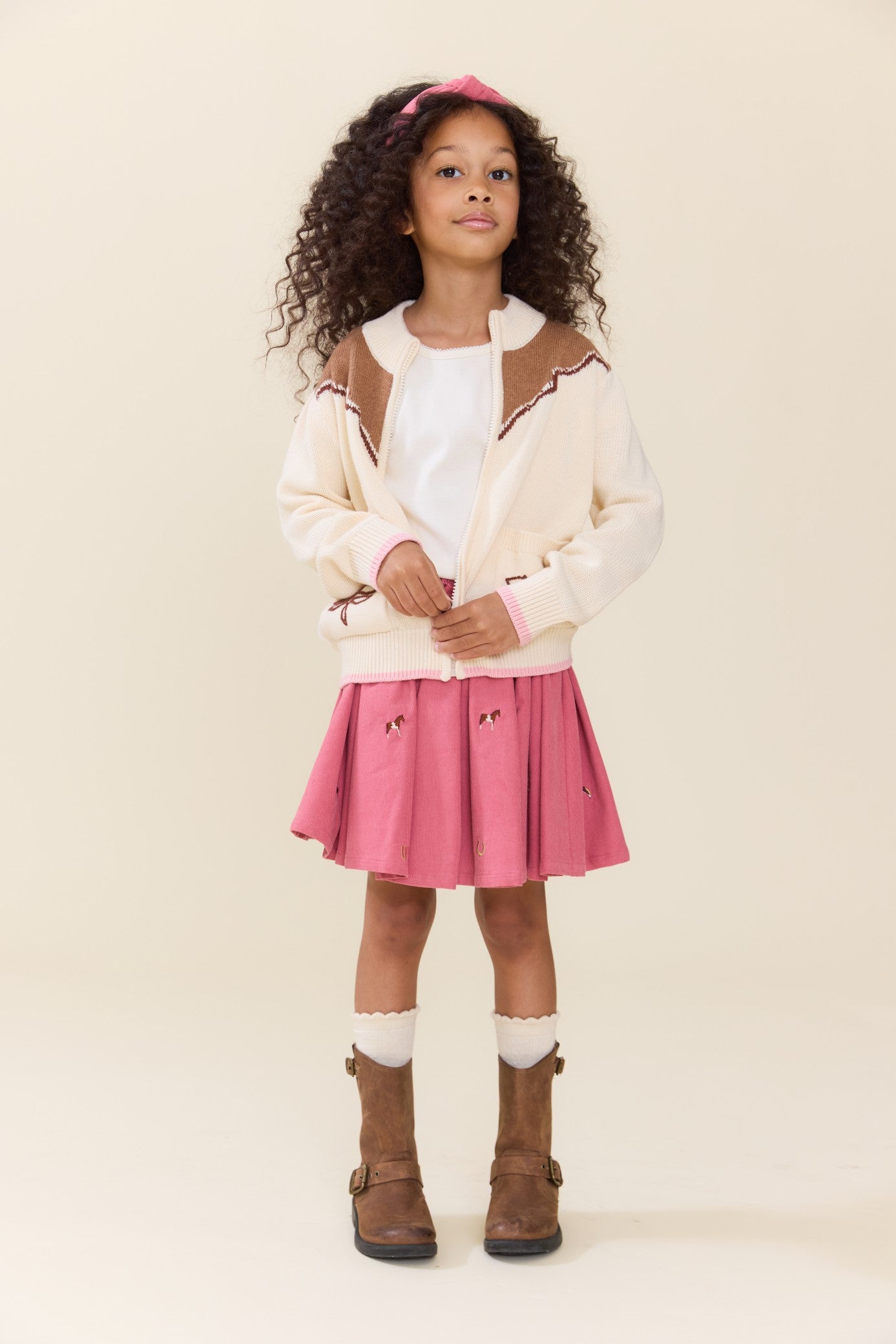 Young girl wearing a pink pleated skirt with horse embroidery styled with a cream cardigan and brown boots from Pink Chicken