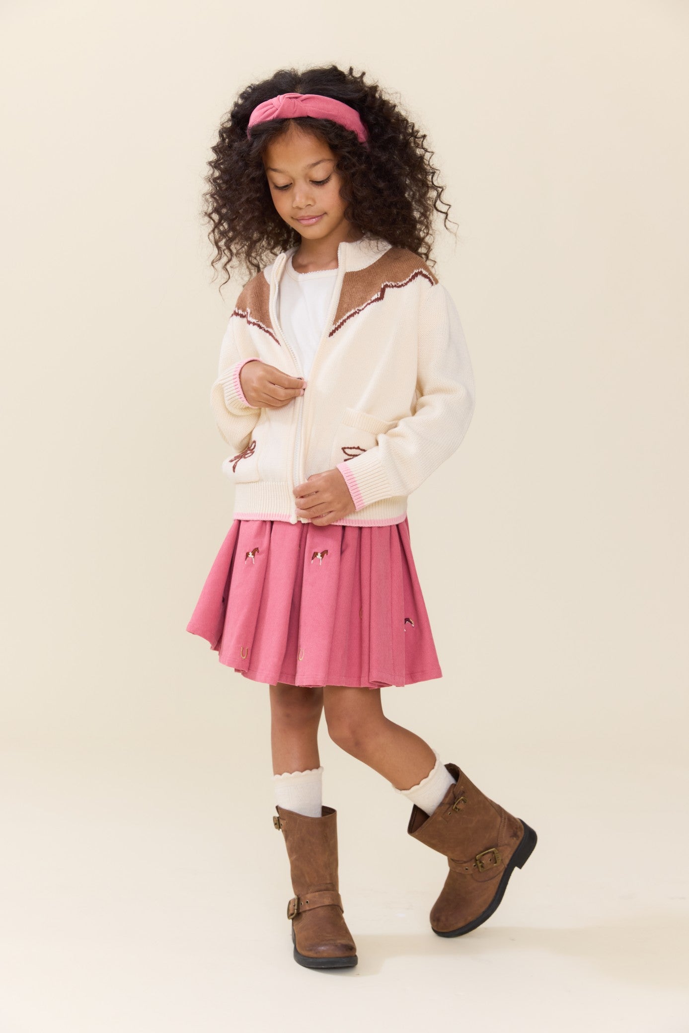 Young girl wearing a pink pleated skirt with horse embroidery a cream cardigan brown boots and a pink headband in a studio setting for Pink Chicken