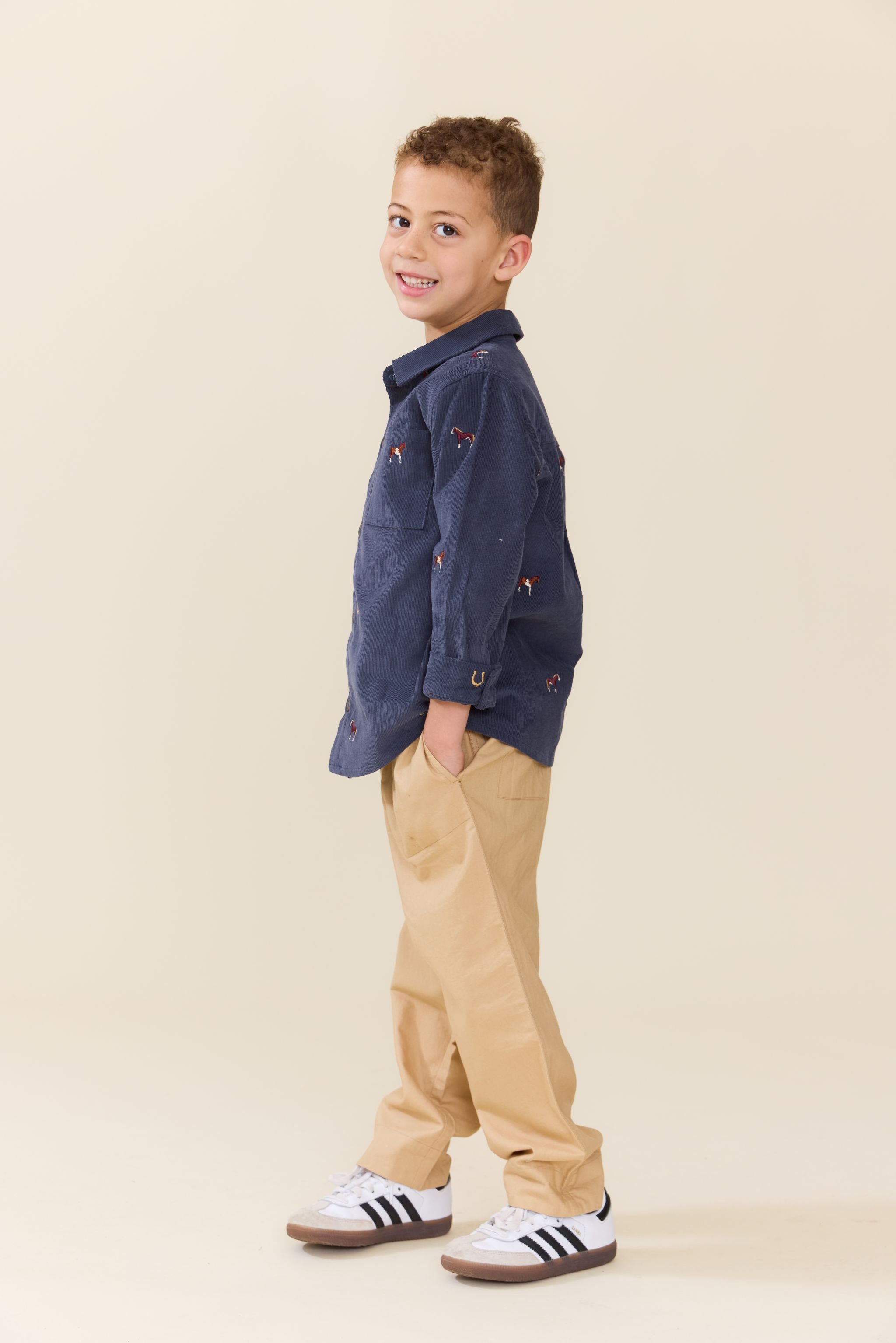 A young boy standing in profile wearing a navy blue corduroy shirt with small embroidered details, beige trousers, and white sneakers with black stripes, against a solid light beige background.
