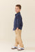 A young boy standing in profile wearing a navy blue corduroy shirt with small embroidered details, beige trousers, and white sneakers with black stripes, against a solid light beige background.