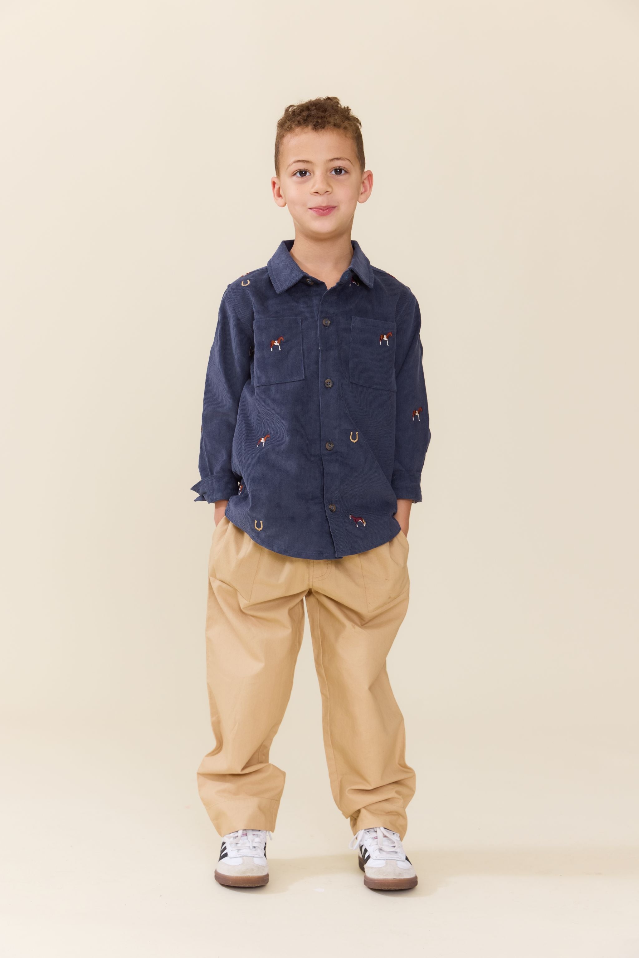 A young boy stands against a solid beige studio background wearing a navy blue long-sleeve button-up shirt with small embroidered details, beige pants, and white sneakers.