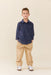 A young boy stands against a solid beige studio background wearing a navy blue long-sleeve button-up shirt with small embroidered details, beige pants, and white sneakers.