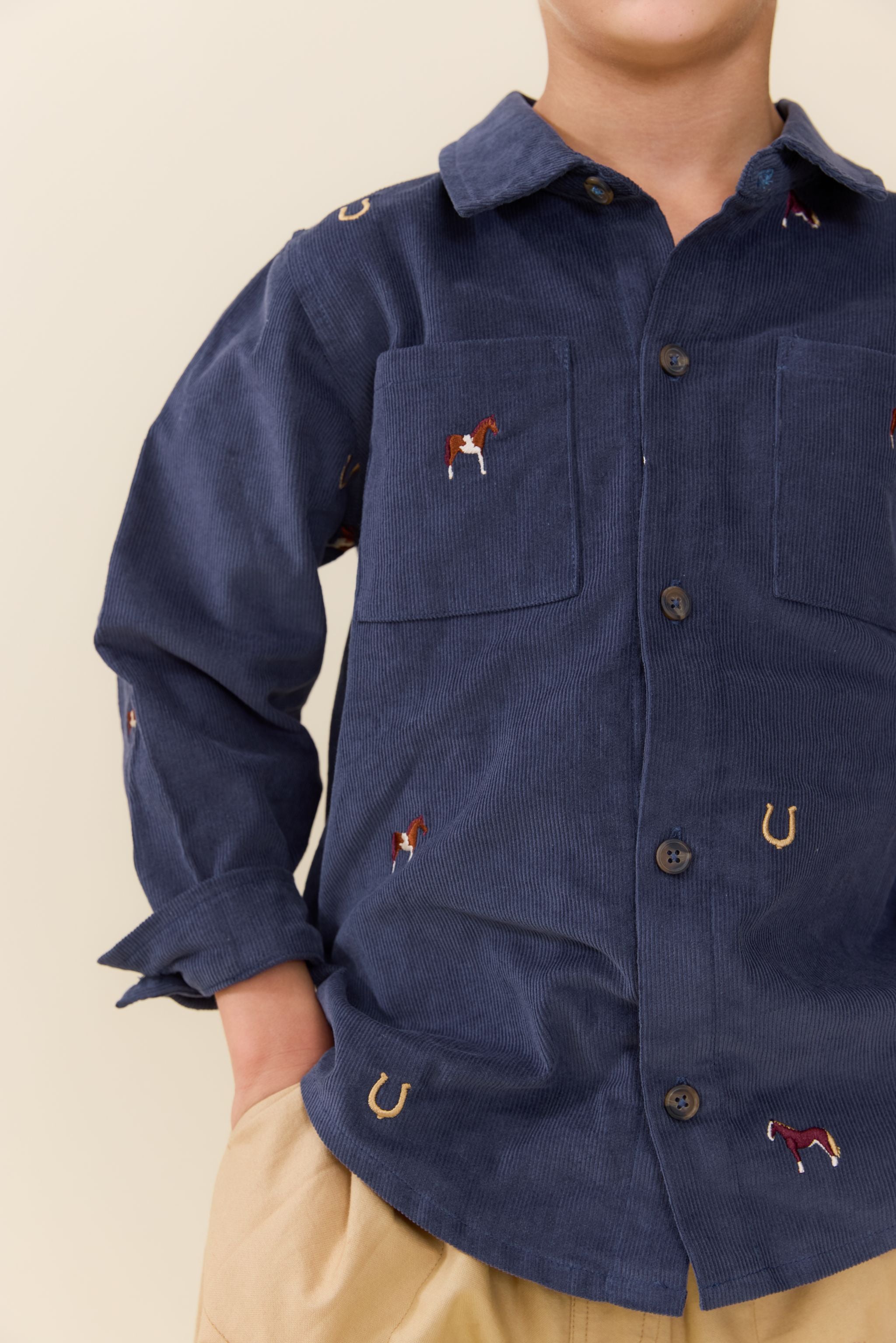 A close-up of a child wearing a navy blue corduroy button-up shirt with embroidered horse and horseshoe motifs, paired with beige trousers, against a solid light beige background.