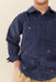 A close-up of a child wearing a navy blue corduroy button-up shirt with embroidered horse and horseshoe motifs, paired with beige trousers, against a solid light beige background.