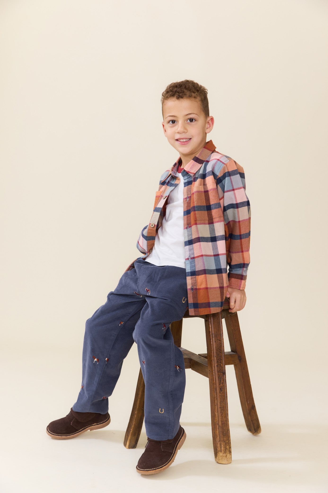 Young boy wearing a brown and blue plaid button down shirt over a white tee with navy pants and brown shoes while seated on a wooden stool by Pink Chicken
