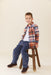 Young boy wearing a brown and blue plaid button down shirt over a white tee with navy pants and brown shoes while seated on a wooden stool by Pink Chicken