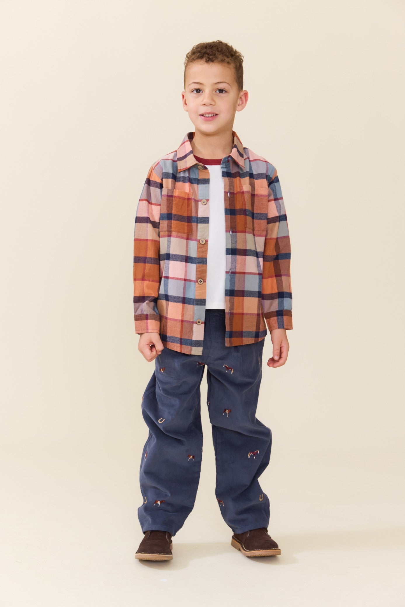 Young boy wearing a brown and multicolor plaid button down shirt with dark blue pants and brown shoes in a studio setting from Pink Chicken