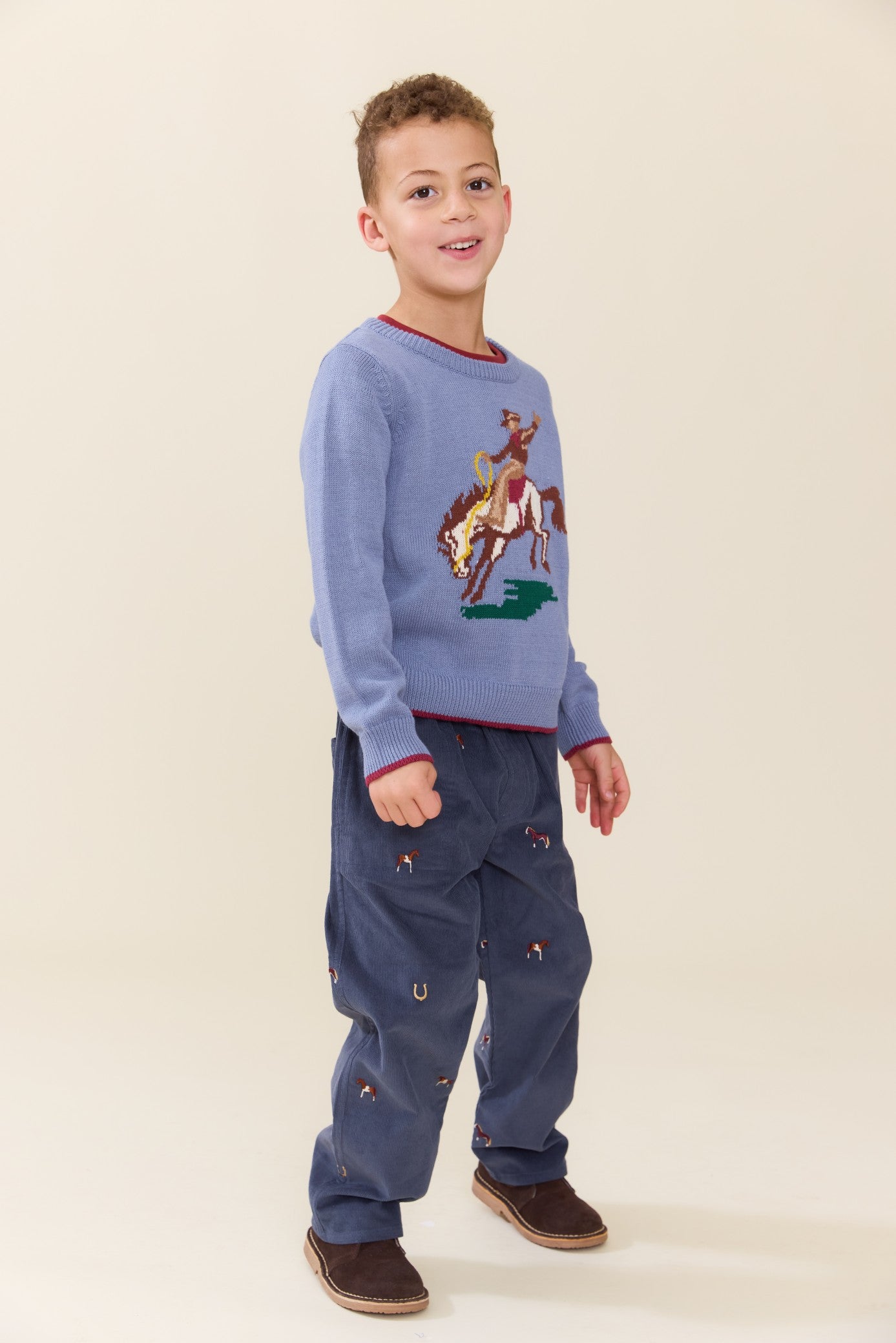 Young boy wearing a blue Pink Chicken sweater with a rodeo horse design and navy pants with western details in a studio setting