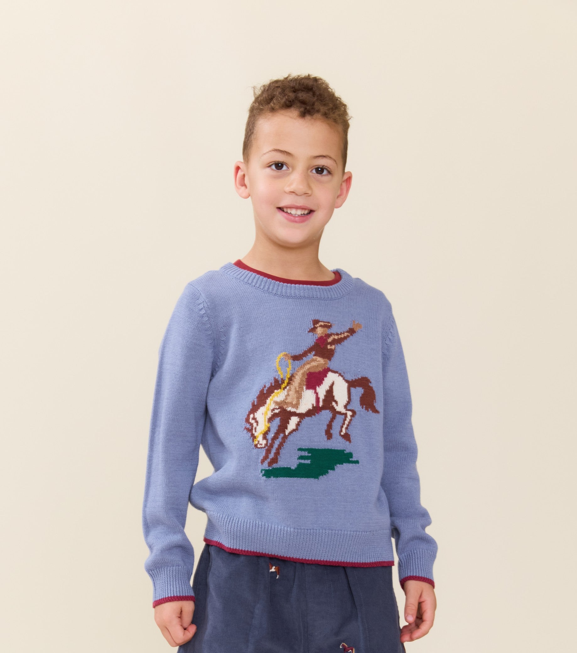 Young boy wearing a blue Pink Chicken sweater featuring a cowboy riding a bucking horse motif with red trim and dark pants