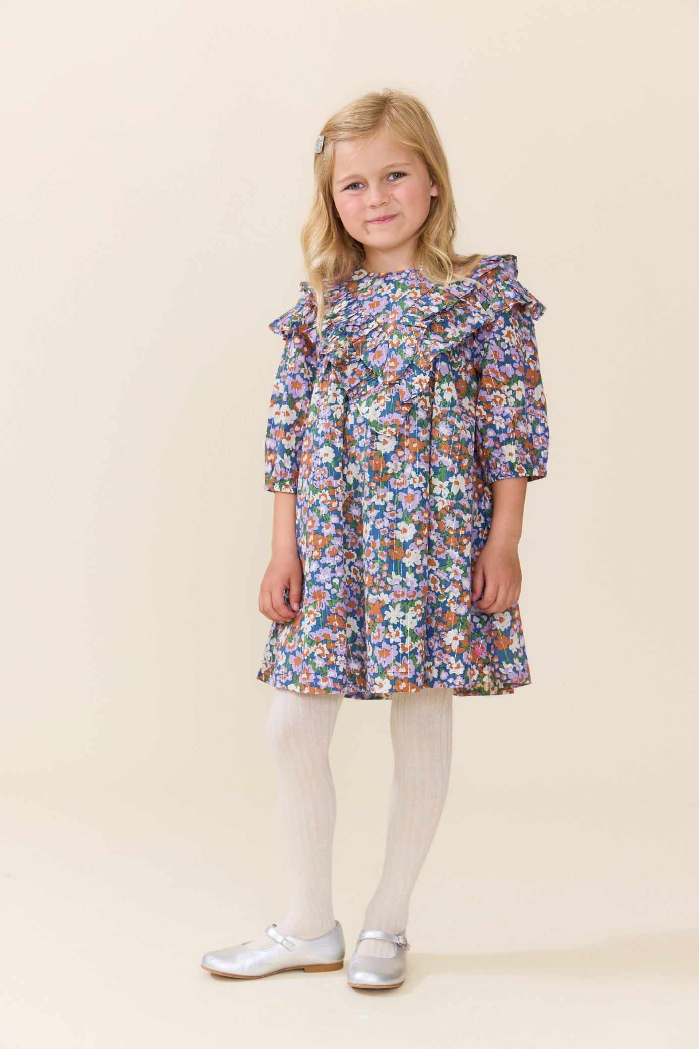 Young girl wearing a blue floral Pink Chicken dress with ruffled shoulders and long sleeves styled with white tights and shoes on a neutral studio background