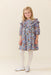 Young girl wearing a blue floral Pink Chicken dress with ruffled shoulders and long sleeves styled with white tights and shoes on a neutral studio background