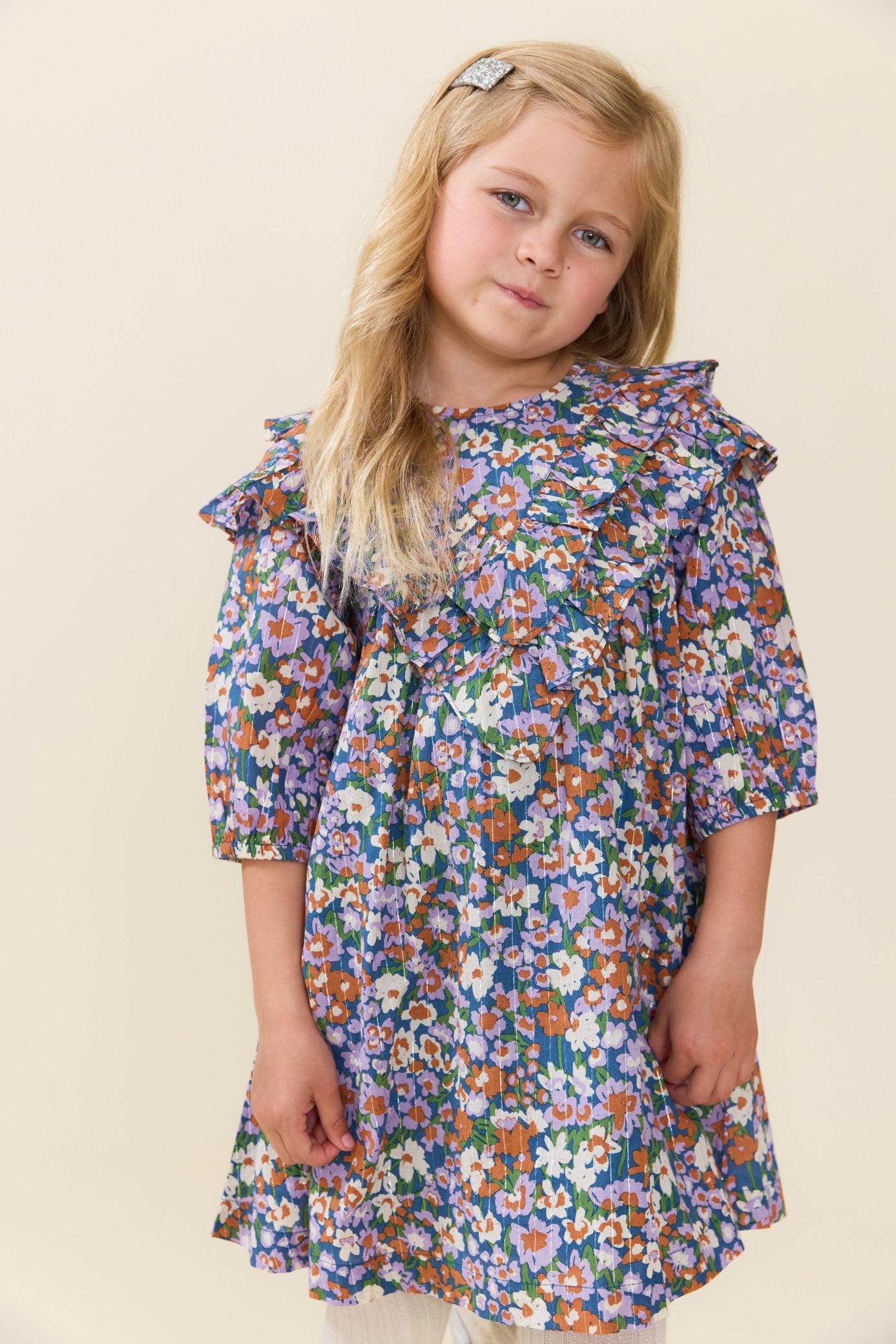 Young girl modeling a blue floral Everly dress by Pink Chicken with three quarter sleeves and a softly flared skirt against a light neutral backdrop