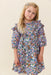 Young girl modeling a blue floral Everly dress by Pink Chicken with three quarter sleeves and a softly flared skirt against a light neutral backdrop