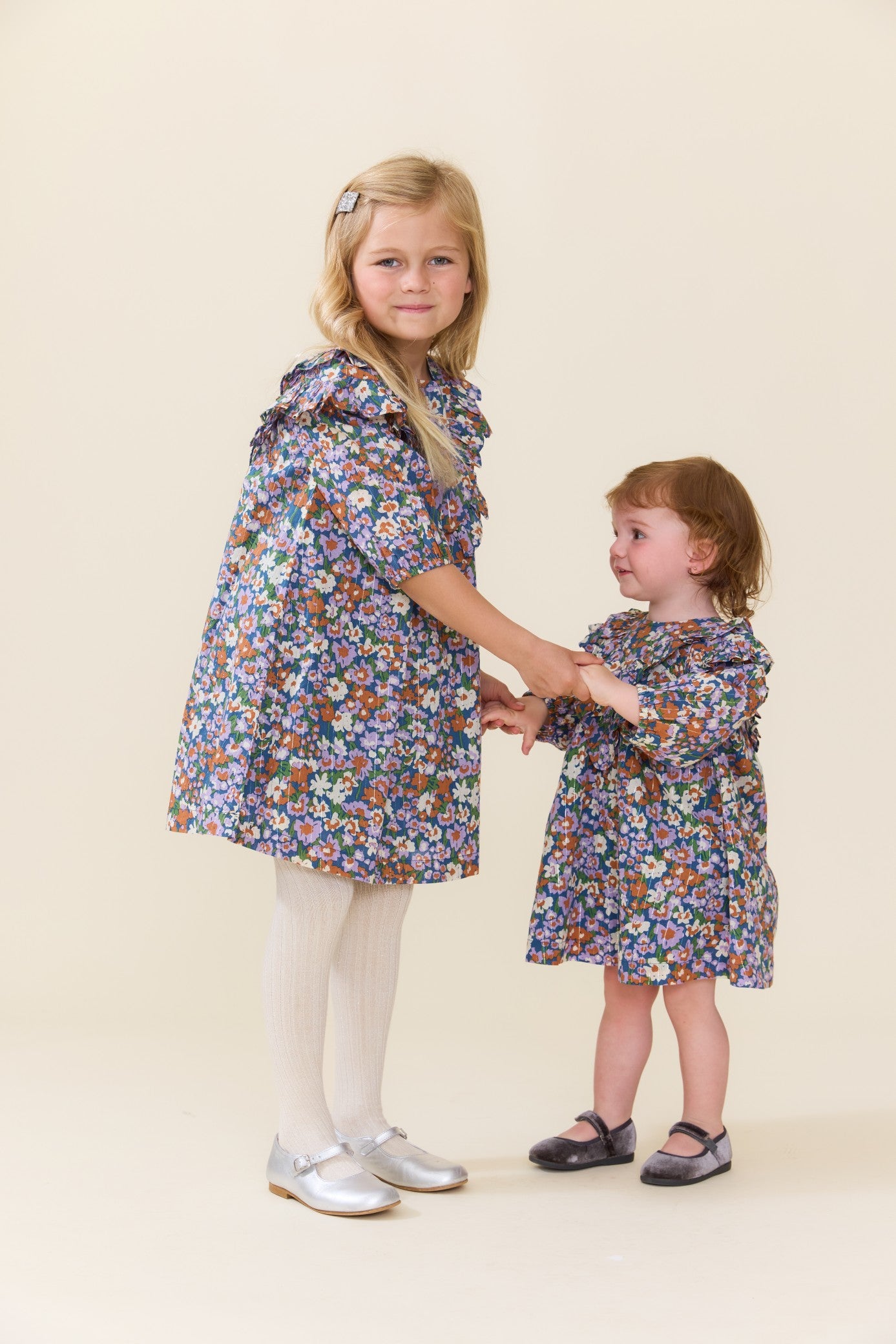 Two girls wearing matching blue floral dresses with white tights and Mary Jane shoes as the older child holds the younger child’s hands Pink Chicken clothing outfit display