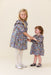 Two girls wearing matching blue floral dresses with white tights and Mary Jane shoes as the older child holds the younger child’s hands Pink Chicken clothing outfit display