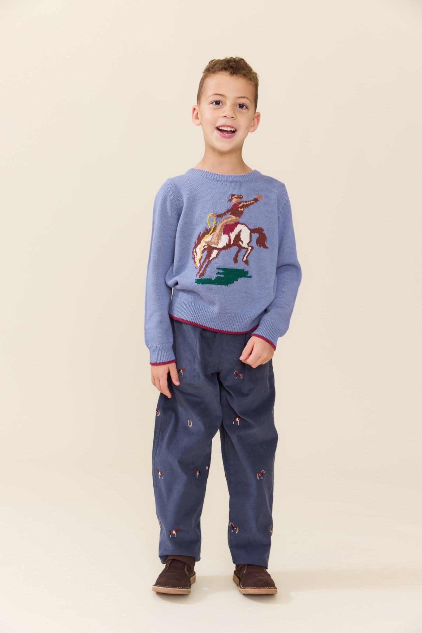 Young boy wearing a blue Pink Chicken sweater with a rodeo horse design and dark patterned pants while standing indoors