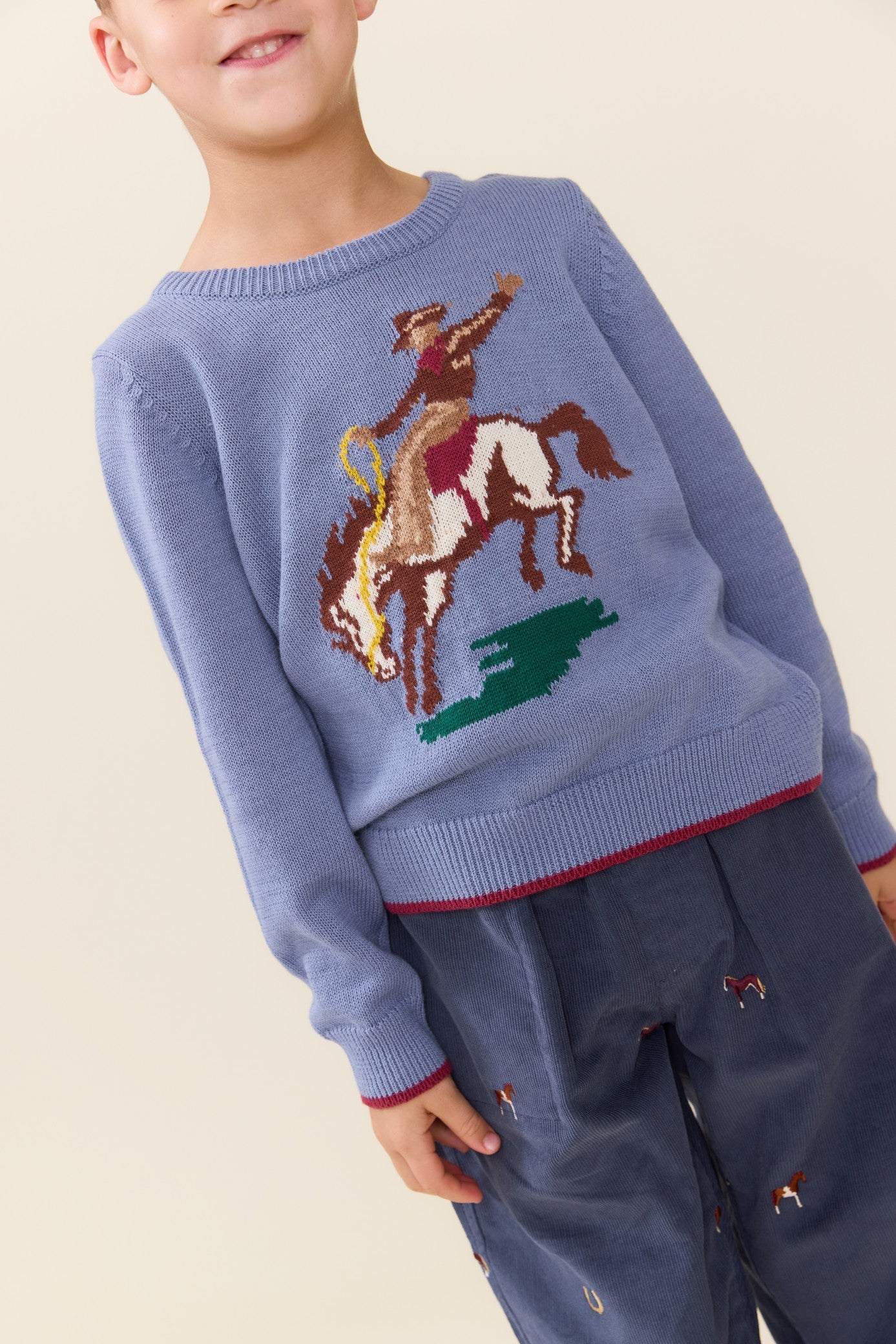 Child wearing a blue Pink Chicken sweater featuring a colorful bucking bronco design with red trim at the cuffs and hem