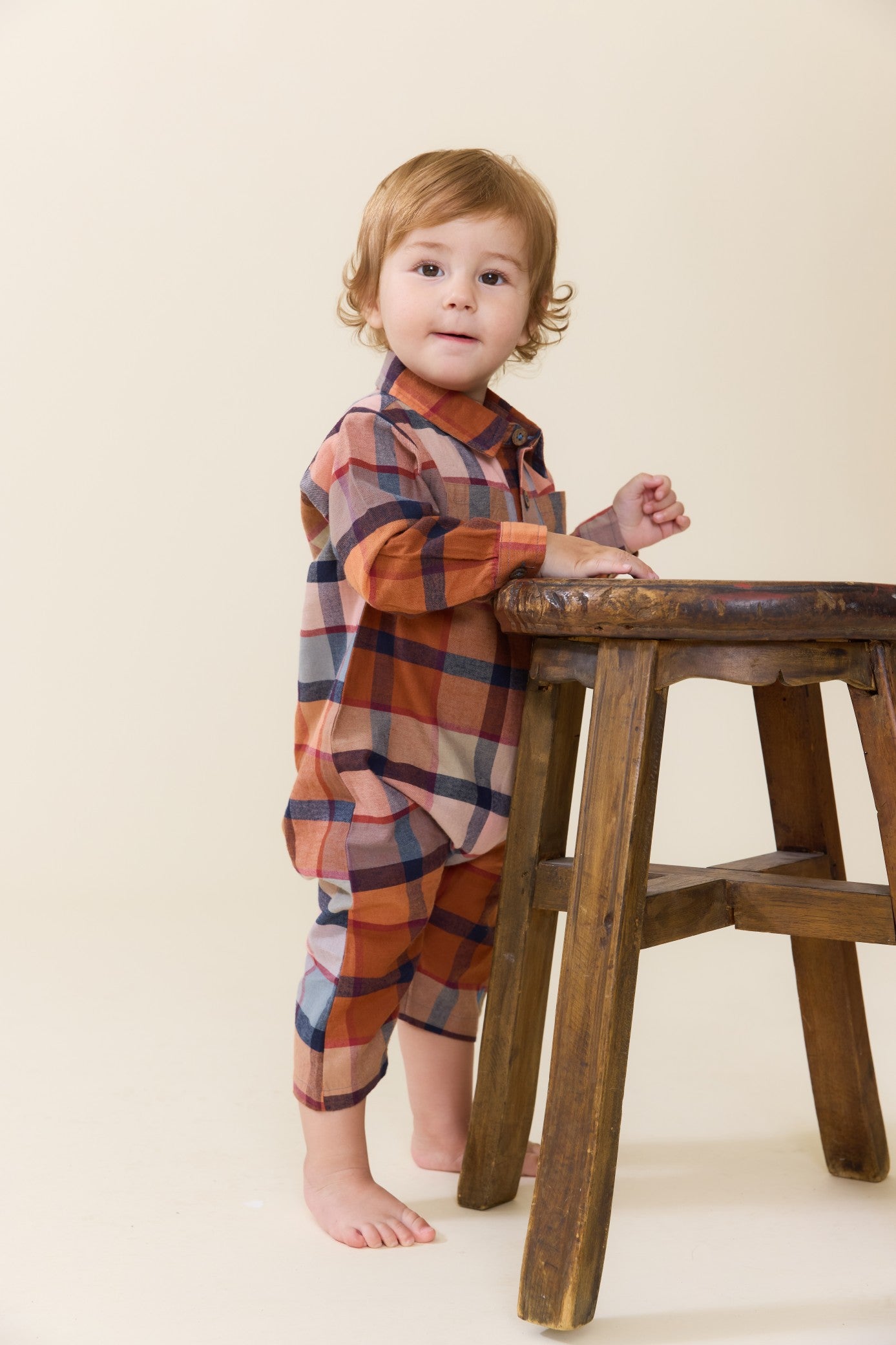 Toddler wearing a brown and rust plaid jumper from Pink Chicken while standing beside a rustic wooden stool