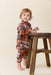 Toddler wearing a brown and rust plaid jumper from Pink Chicken while standing beside a rustic wooden stool