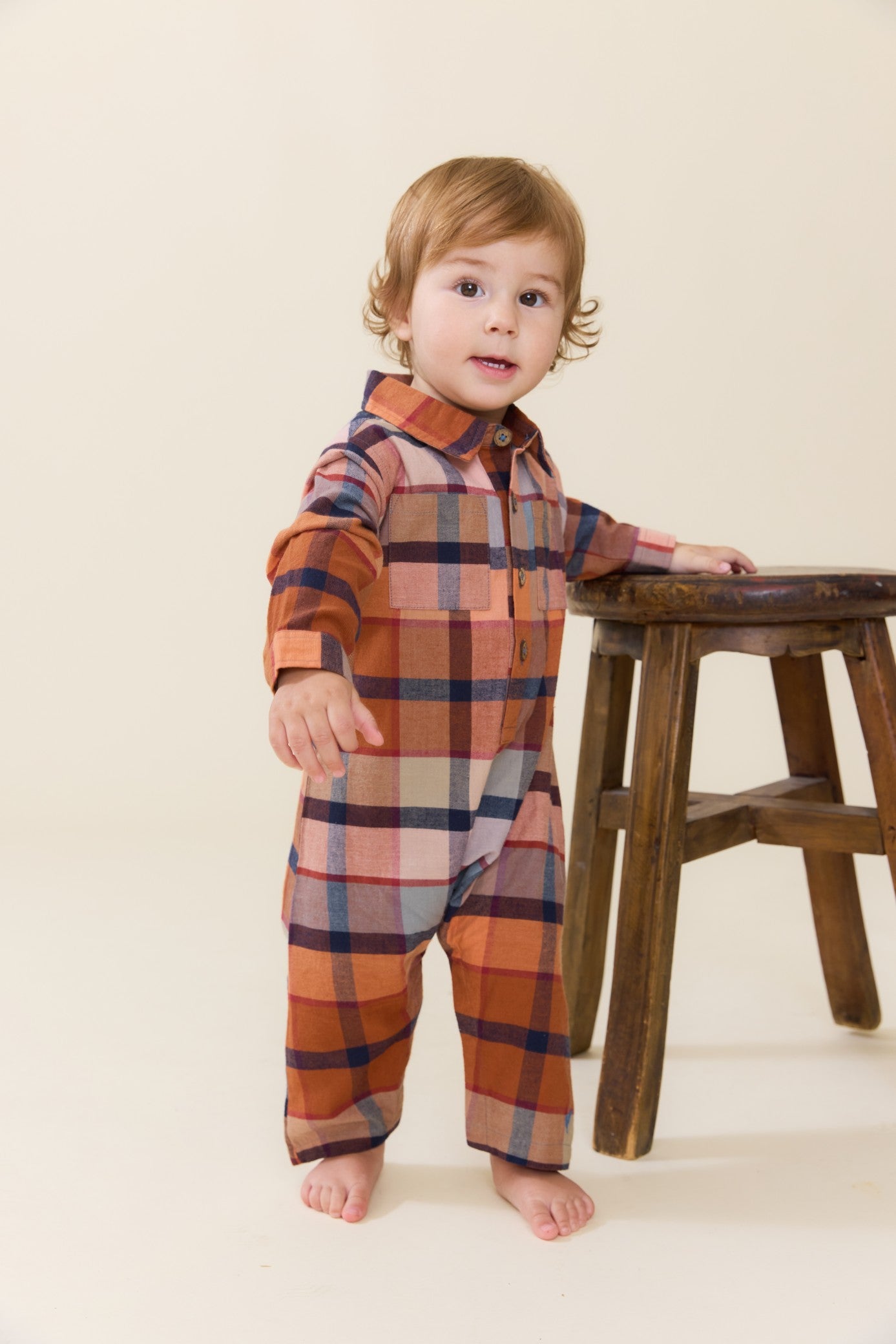 Toddler wearing a brown and orange plaid jumper with long sleeves and button front standing beside a wooden stool from Pink Chicken