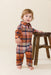Toddler wearing a brown and orange plaid jumper with long sleeves and button front standing beside a wooden stool from Pink Chicken