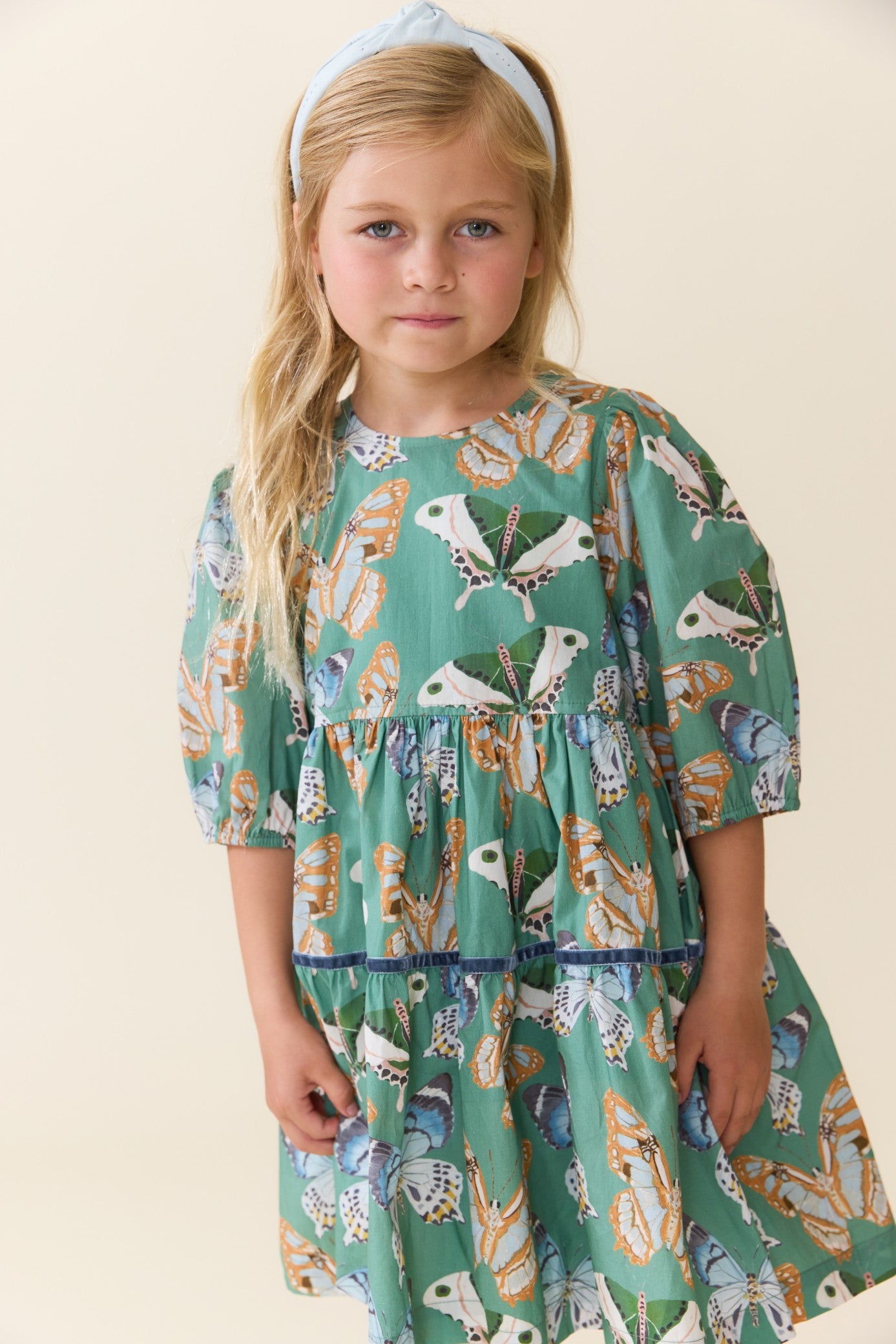 Young girl wearing a green butterfly print dress by Pink Chicken with a gathered waist three quarter sleeves and a flowing skirt