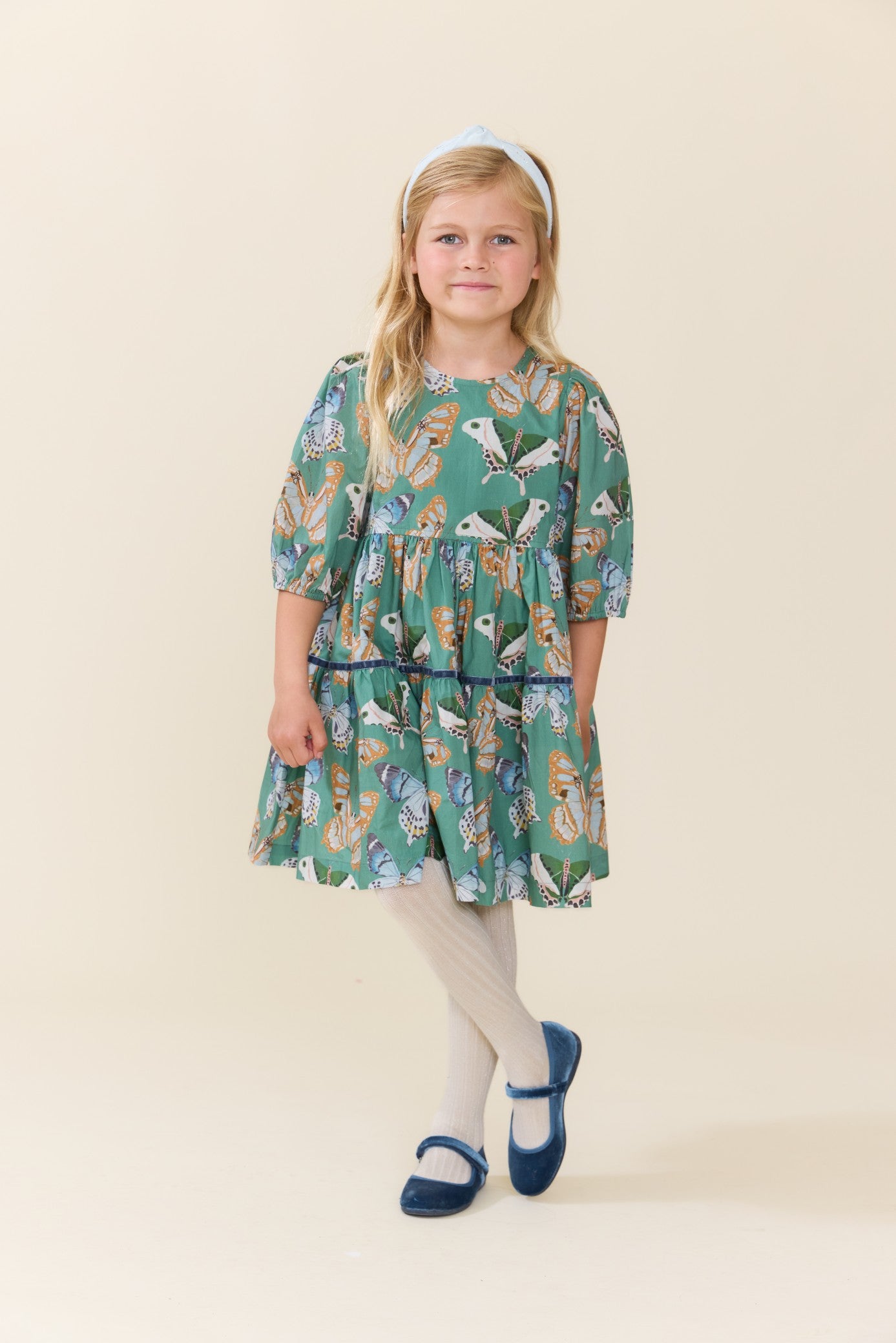 Young girl wearing a green butterfly print dress with a gathered waist and puff sleeves paired with white tights and navy Mary Jane shoes from Pink Chicken