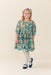 Young girl wearing a green butterfly print dress with a gathered waist and puff sleeves paired with white tights and navy Mary Jane shoes from Pink Chicken