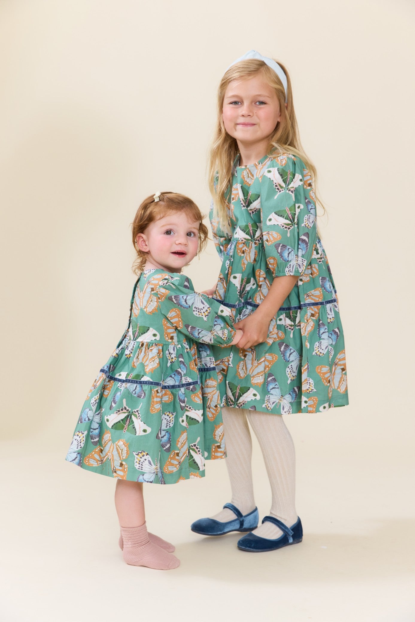 Two girls wearing matching green butterfly print dresses with tights and Mary Jane shoes from Pink Chicken in a charming studio portrait