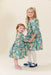 Two girls wearing matching green butterfly print dresses with tights and Mary Jane shoes from Pink Chicken in a charming studio portrait