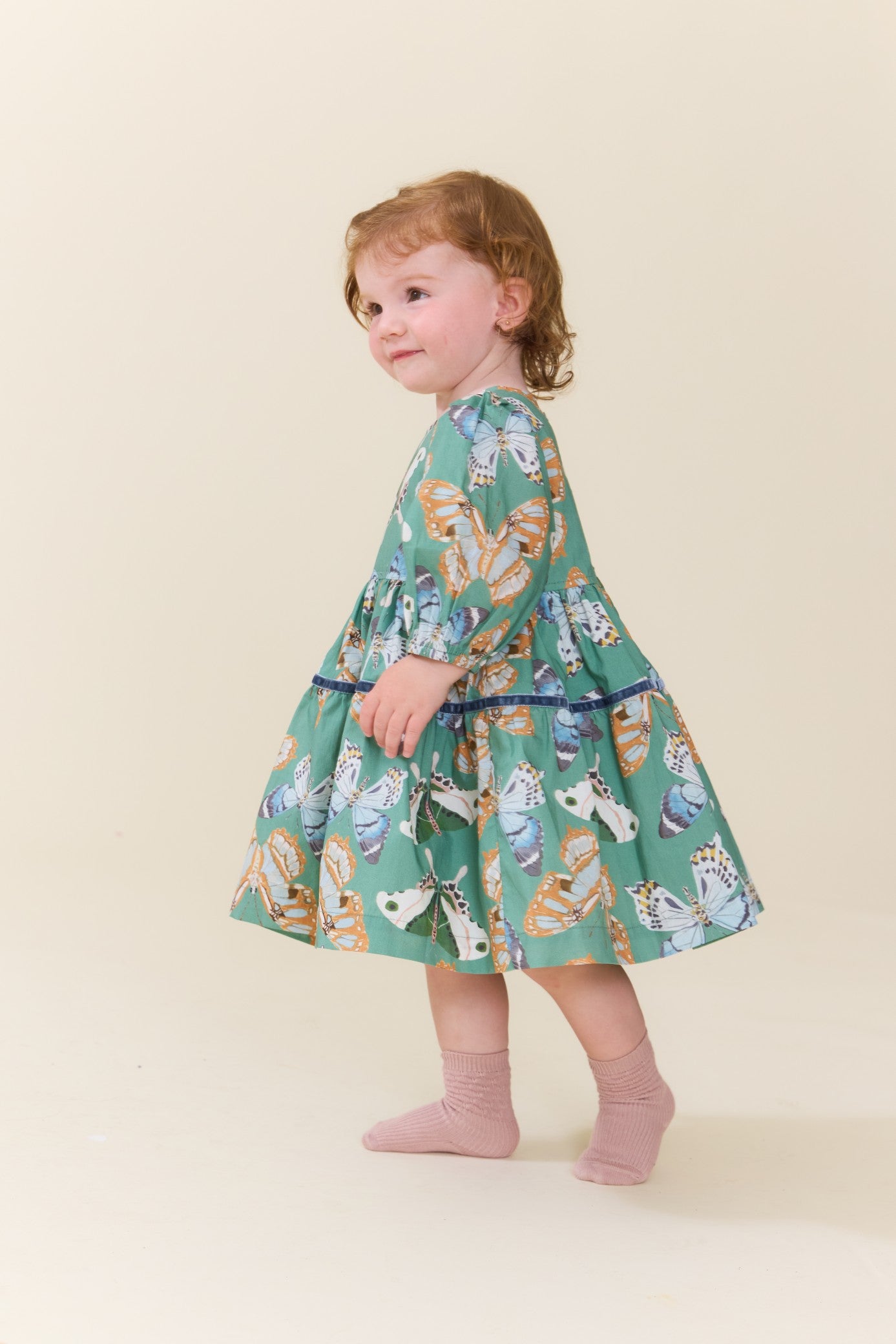 Young girl modeling a green butterfly print dress with long sleeves and a full skirt while wearing pink socks in a studio from Pink Chicken