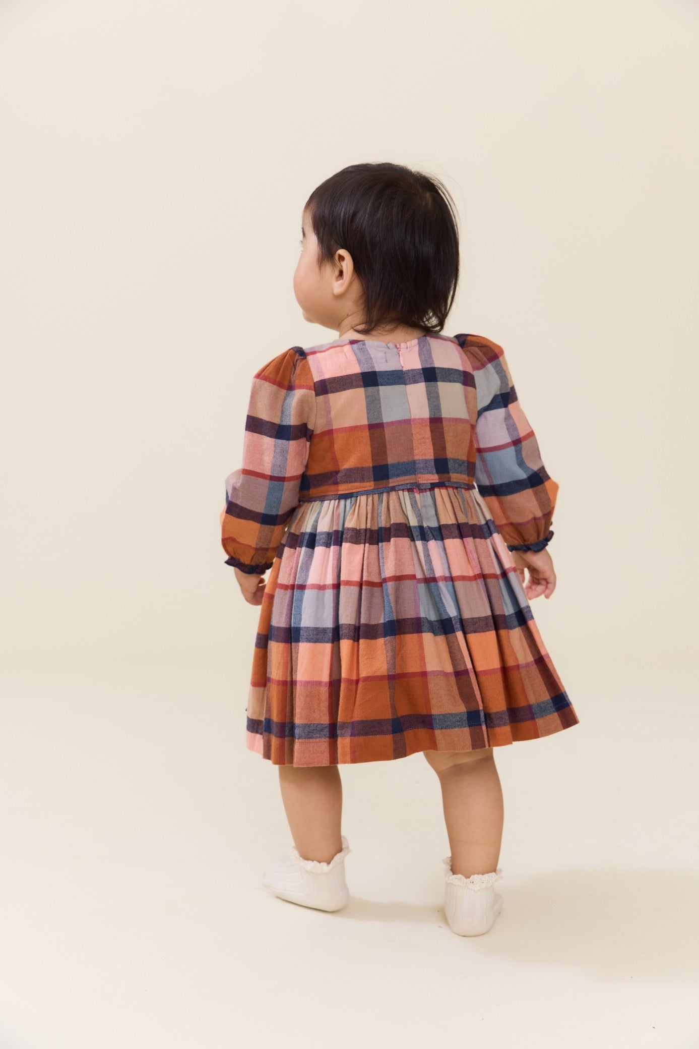 Toddler girl wearing a long sleeve plaid dress with a gathered skirt and white shoes from Pink Chicken posed against a neutral background